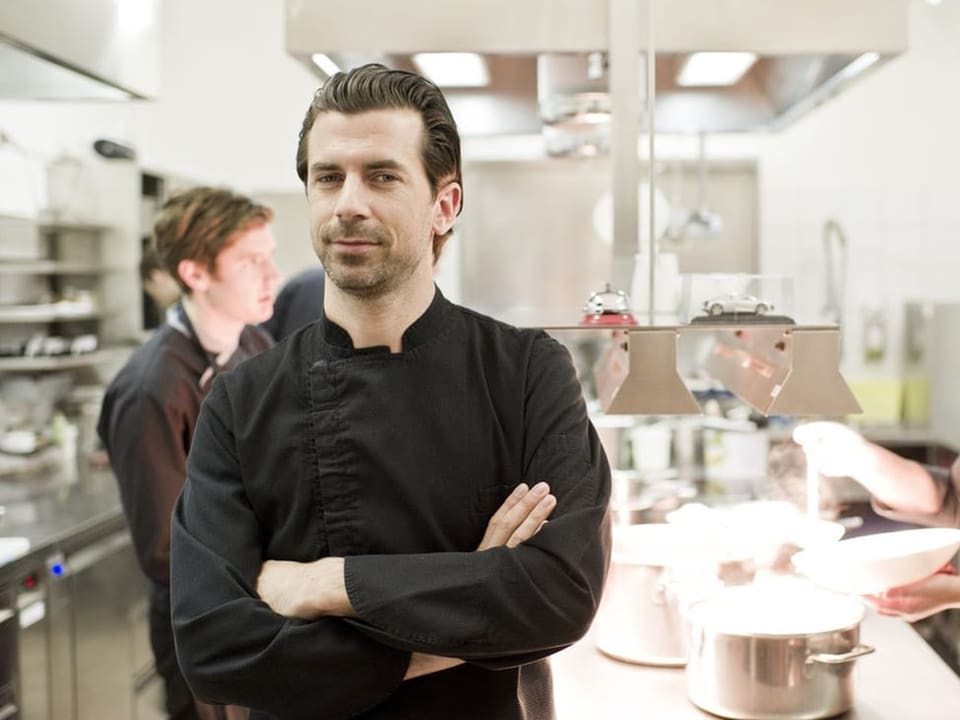 A man stands in a kitchen with his arms crossed.