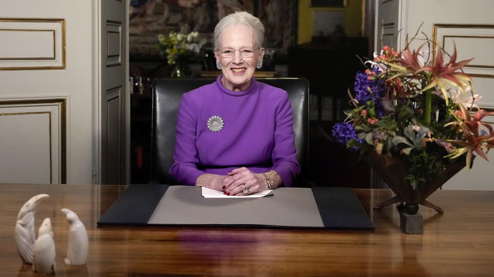 Margrethe II in front of a desk.  To her right is a bouquet of flowers; she is wearing a purple dress.