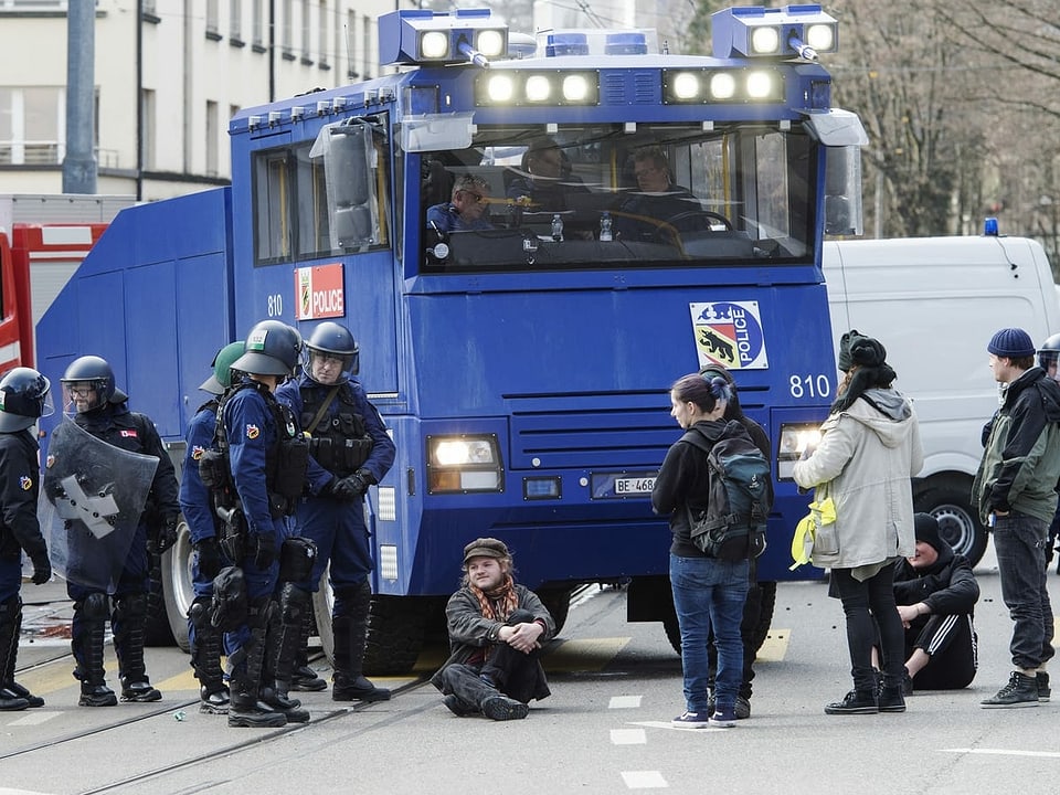 A man sits on the ground in front of a police water cannon.