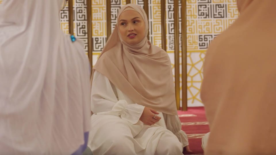 Nina in a white robe and a beige headscarf in the mosque looks at two friends.