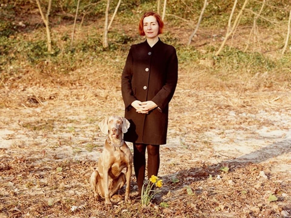 A woman with short, red hair and a black coat stands at the edge of the forest, a dog next to her.