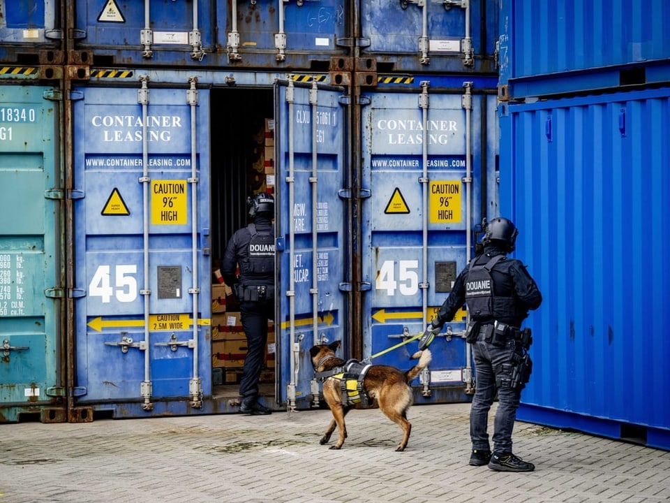 Two police officers search for drugs in a shipping container with a sniffer dog.