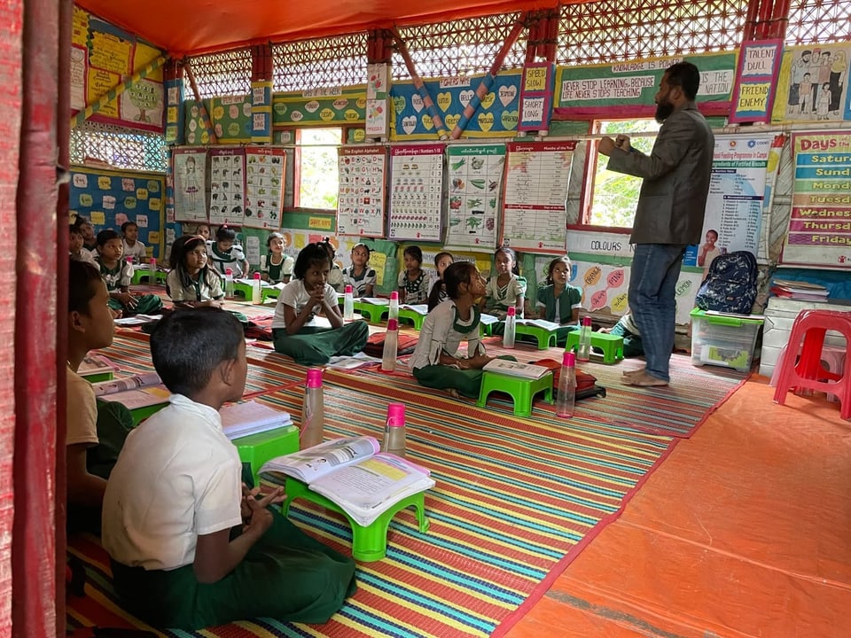Children sit on the floor in a very colorfully decorated school room.  The teacher stands and explains.