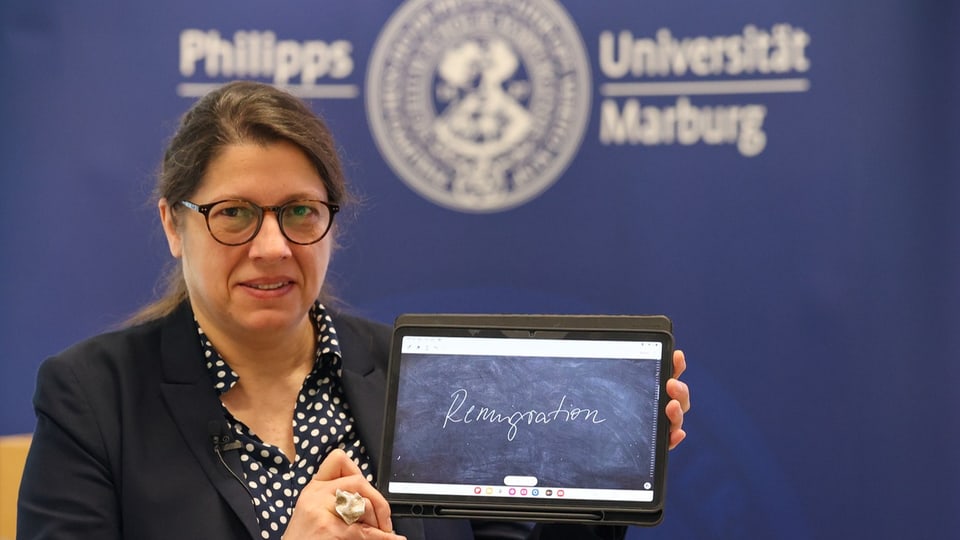 A woman holds an iPad with the word “remigration” written on a blackboard