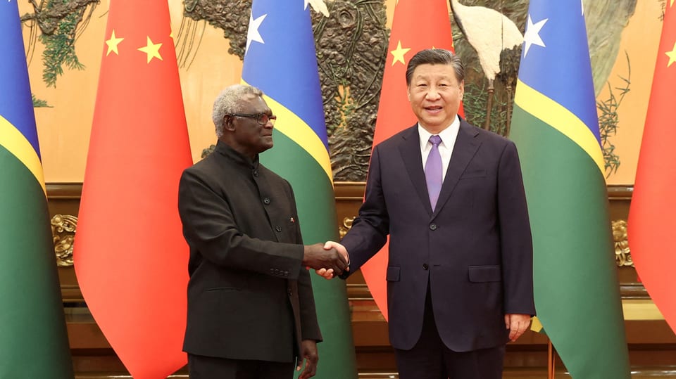 A dark-skinned man and a Chinese man shake hands in front of flags.