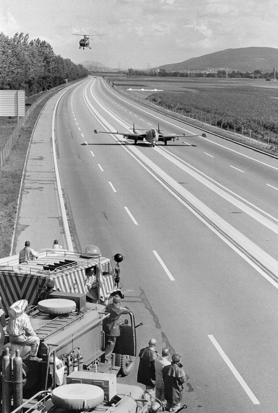 Military jet taxis on a stretch of highway;  vehicles are placed on the side of the road;  A helicopter circles above