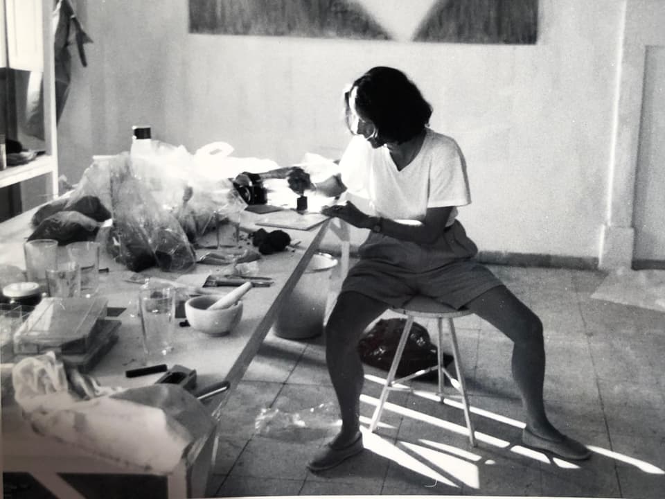 A woman sits at a crowded table in a studio and paints a picture. 