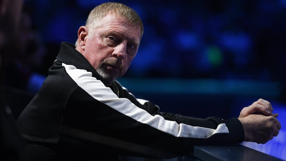 Boris Becker with short hair in a black and white trainer, looking into the camera, leaning on the railing.