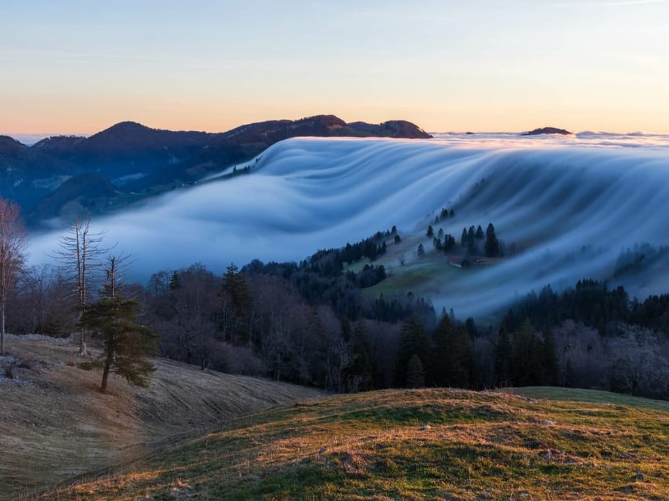 Hilly landscape with fog on the right, which flows like a waterfall to the left and dissipates. 