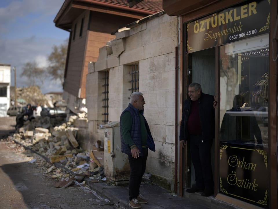 A man talks to a friend at the entrance to a store.  Behind it you can see damage from the earthquake a year ago.