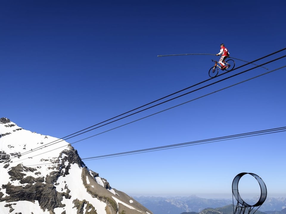 Nock rides a bike over the rope of the cable car.  He holds a long staff for balance.