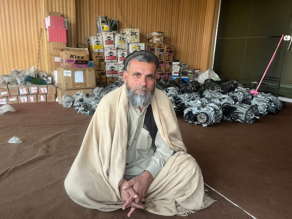 Man in traditional clothing sits on the ground.  Behind it are car parts.