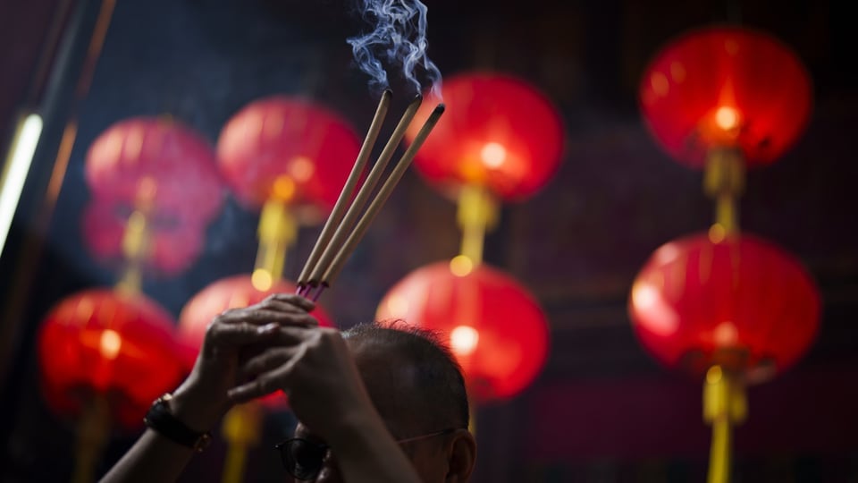 A man holds incense sticks in the air, red lanterns in the background