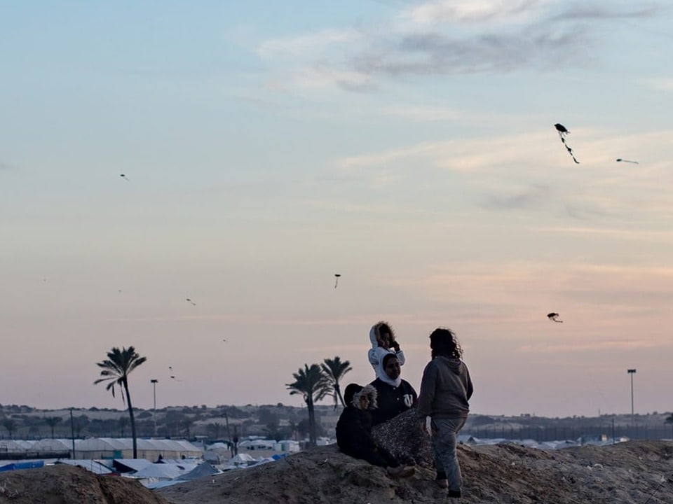 Children fly kites, behind them is a refugee camp