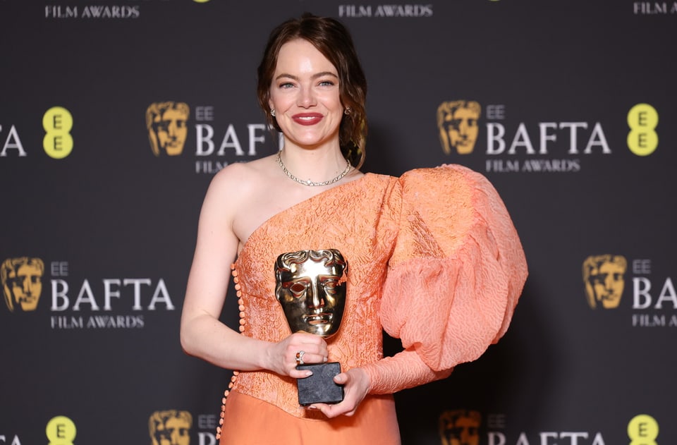 Emma Stones stands in front of the Bafta wall and holds the trophy in her hand.