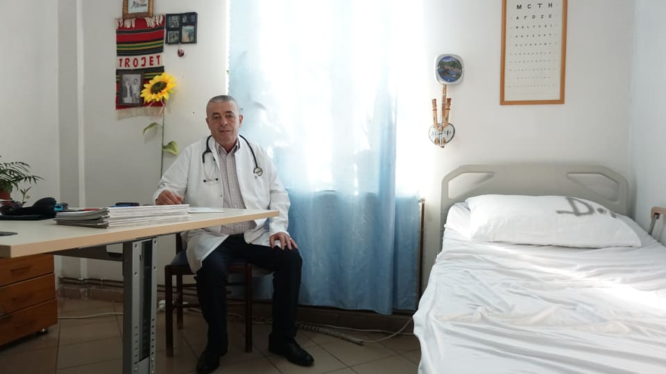 A man in a white coat sits at a desk next to a hospital bed