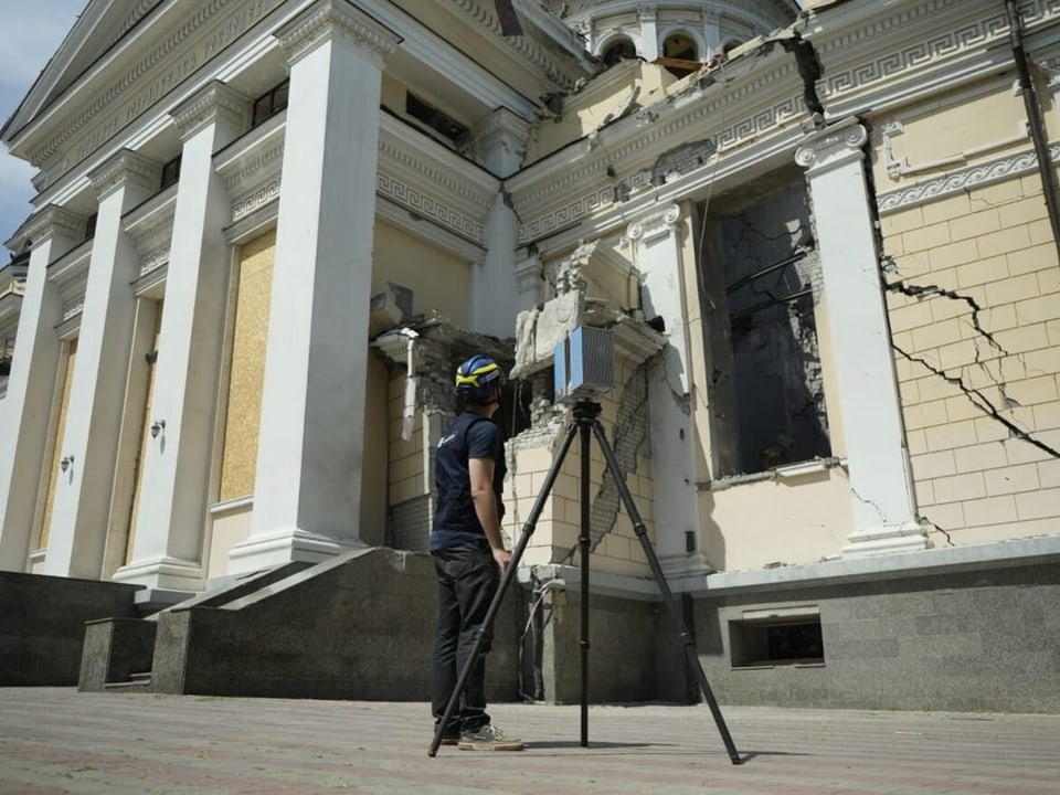 A yellow and white facade with cracks, in front of it a man in a helmet, in front of him a device with a tripod.
