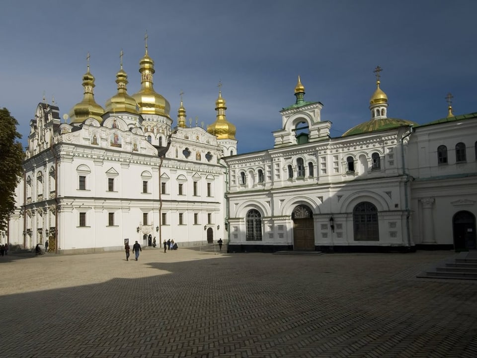 Monastery with golden onion domes, a large square in front of it, only a few people.  The facade of the monastery is white.