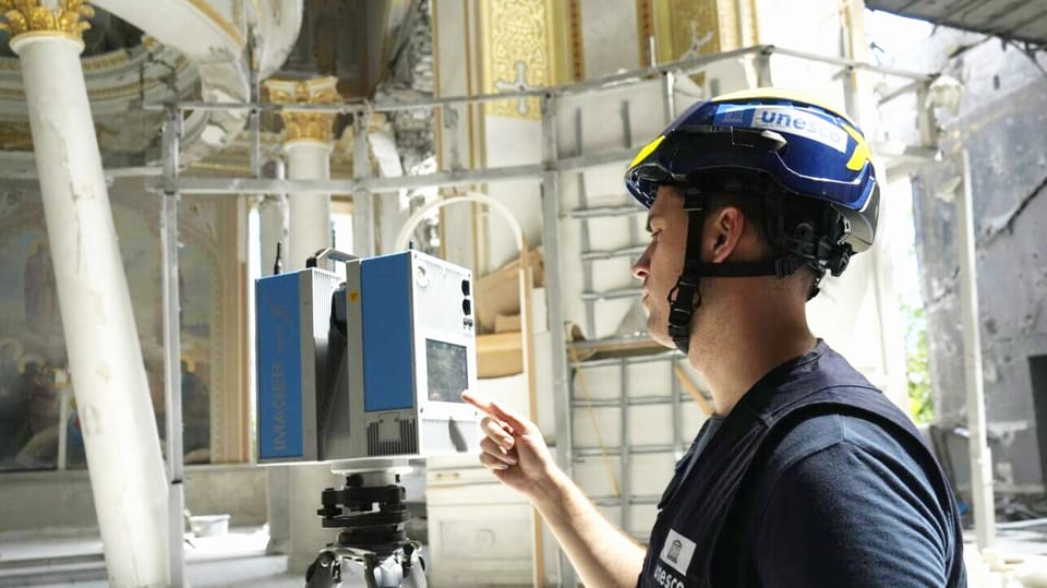 A young man wearing a blue T-shirt and a helmet with “Unesco” written on it.  He operates a device on a tripod.