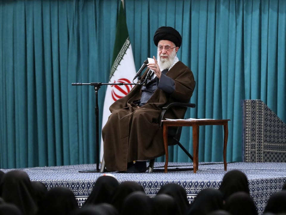 Ali Khamenei sits on a stage and speaks into microphones.  Heads of audience members can be seen in front of the stage.