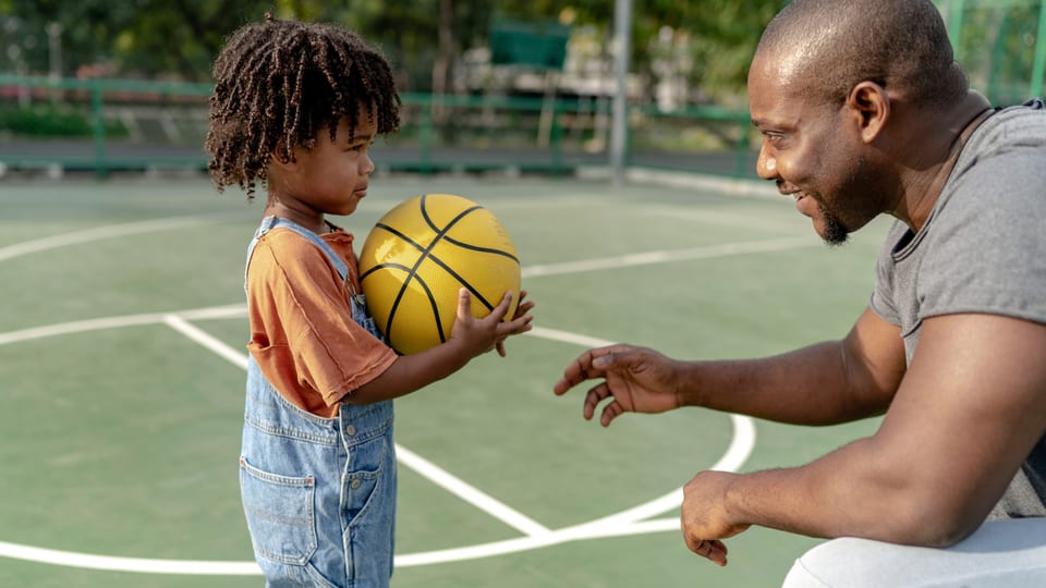 Kind (links) mit gelbem Basketball, Vater rechts, kniet zu ihm runter.