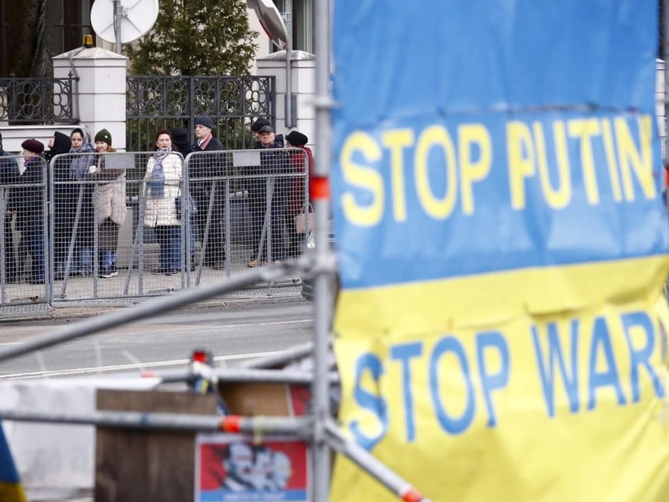 In the background: queue of people.  In the foreground Ukrainian flag with the inscription “Stop Putin – Stop War”