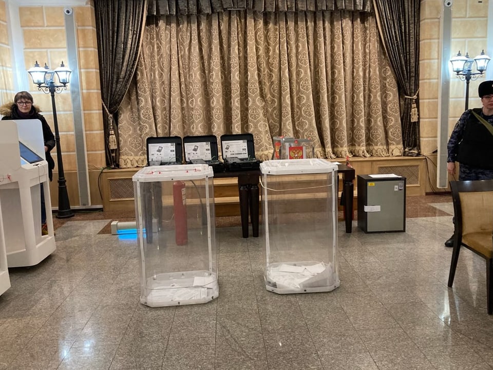View of two transparent ballot boxes;  two people are standing next to it.