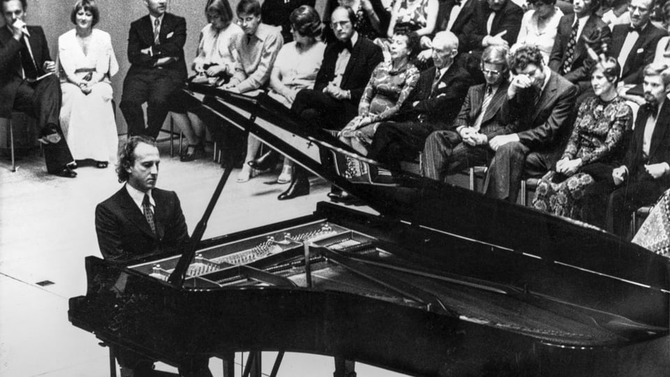 Black and white photo: A man sits at the piano, his audience sits behind him (take).