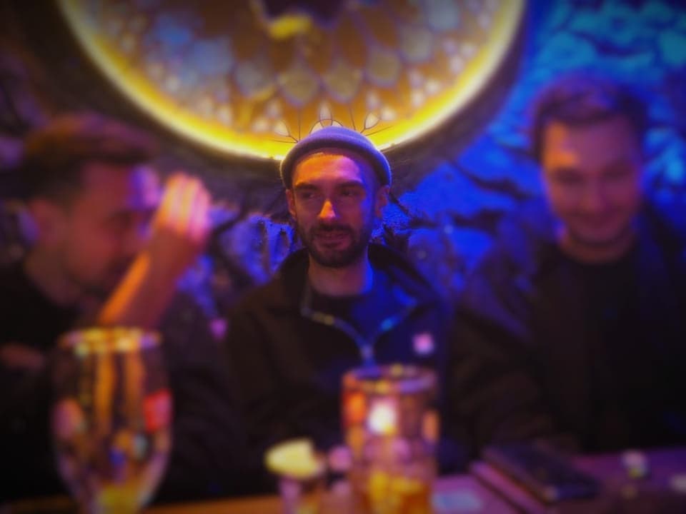 Junger Mann mit Mütze, sitzt zwischen zwei Männern am Tisch in einer Bar.