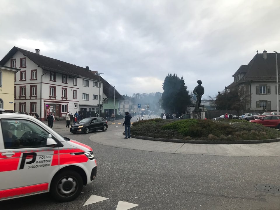 A cloud of smoke hangs over a street.  There is a police car and people on the street.