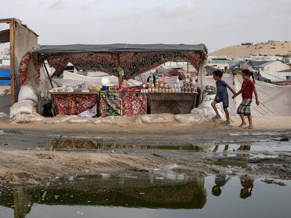 Children walk past a market stall in a refugee camp with reflection in the water.